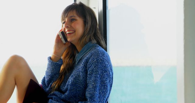 Young woman smiling and talking on smartphone near window - Download Free Stock Images Pikwizard.com