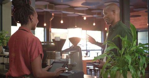 Barista serving customer using tablet and phone at sunlit coffee counter with grinders