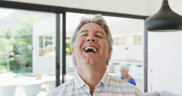 Senior man shown laughing in a modern home with large windows and bright lighting. Suitable for advertisements related to happiness, senior lifestyle, modern living, or healthcare. Can also be used for articles on aging positively, mental health, or living spaces.