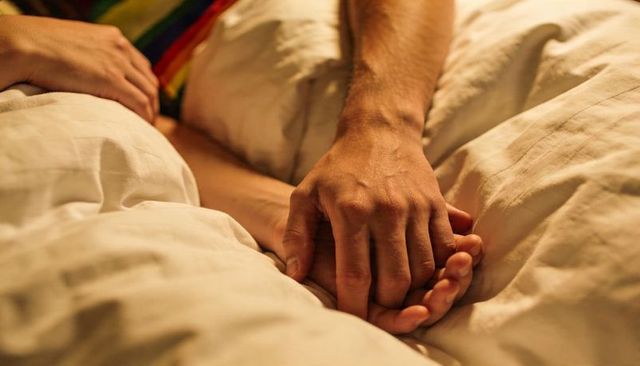 Holding interlaced hands on cream duvet under warm golden light conveying tenderness and cozy intima