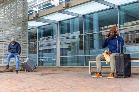 Urban travelers waiting at glass terminal, black man calling and eating with luggage and reflections