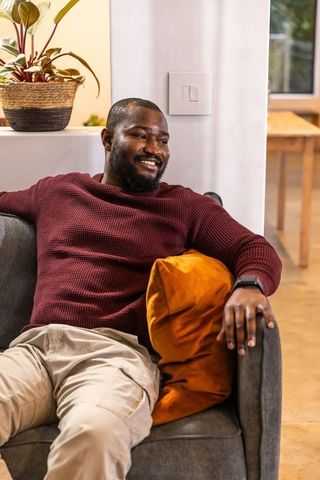 Relaxing african american man reclining on gray sofa smiling with orange pillow in cozy living room