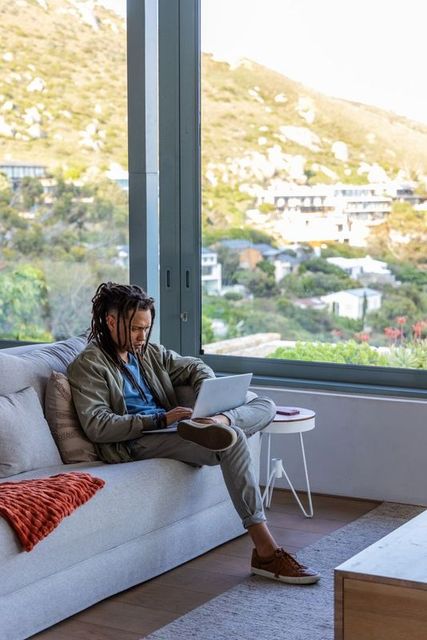 African american man working remotely on laptop while lounging on modern living room sofa