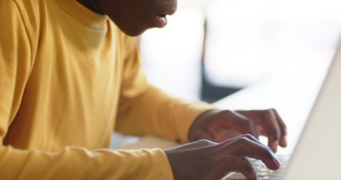 African youth typing on laptop in bright workspace wearing yellow long-sleeve shirt