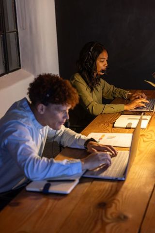 Evening customer support team collaborating at wooden desk with headsets and laptops