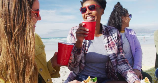 Group of friends relaxing at beach on sunny day, holding red cups and laughing. Possible use in advertisements for summer activities, beach products, food and drink brands, or vacation destinations to illustrate fun and leisure.