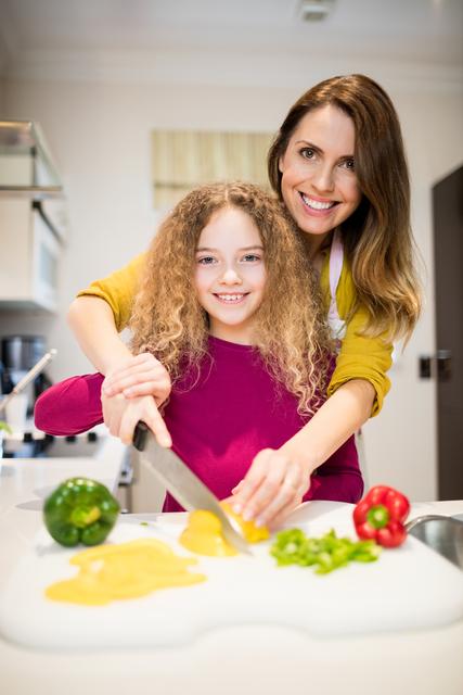 Mother and daughter are preparing a meal together in the kitchen, cutting vegetables. This image can be used for promoting family bonding, healthy eating habits, parenting tips, and lifestyle blogs. It is ideal for advertisements related to kitchenware, cooking classes, and family-oriented products.