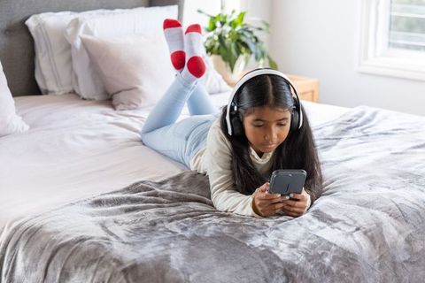 Indian schoolgirl lying on bed listening with headphones and using smartphone at home