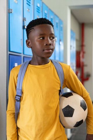 African american boy standing by blue lockers holding soccer ball and backpack