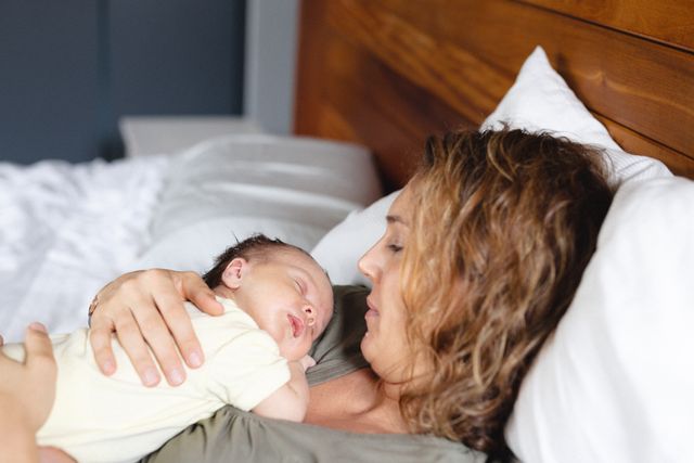 Caring mid adult caucasian mother with cute sleeping newborn baby lying on bed at home, copy space. Unaltered, family, love, togetherness, care, innocence, motherhood, babyhood.