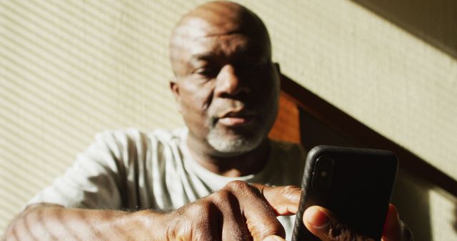 Thoughtful african american senior man sitting at home in the sun using smartphone. retirement lifestyle, spending time alone at home.