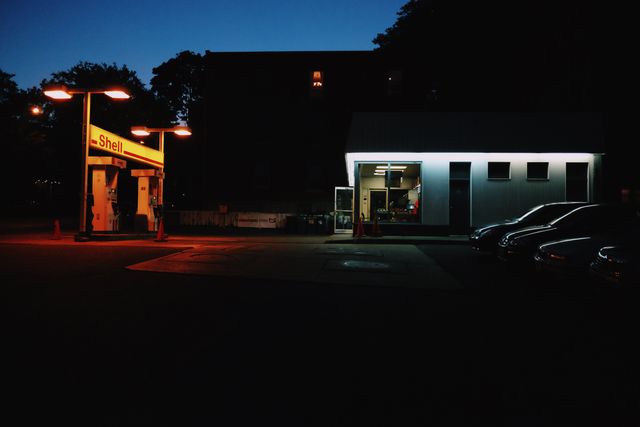 Features gas station illuminated by streetlights at night with convenience store nearby. Ideal for themes of travel, nighttime urban landscapes, city life, solitude, and roadside services.