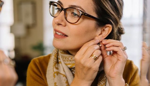 Woman adjusting gold dangle earrings wearing tortoiseshell glasses and patterned scarf