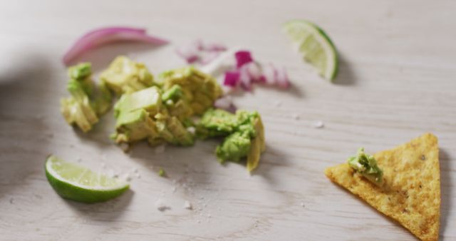 Shows ingredients for making guacamole on a wooden table. Fresh avocado chunks, lime wedges, red onion slices are placed next to a tortilla chip with avocado. Useful for cooking blogs, recipe websites, culinary presentations, healthy eating promotions.