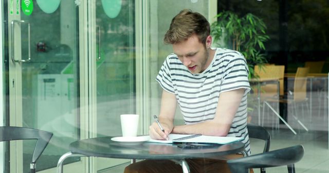 A young Caucasian man is focused on writing in a notebook at an outdoor cafe table, with copy space. His casual attire and concentrated expression suggest he could be a student or a creative professional drafting ideas.