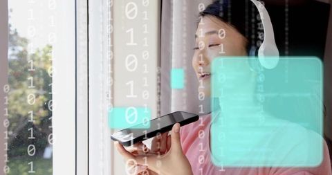 Asian woman speaking to smartphone while wearing headset, holding device by window