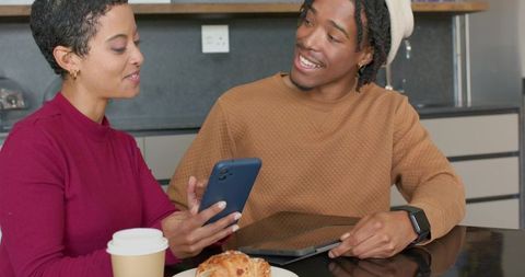 Diverse friends sharing smartphone and tablet while enjoying coffee and pastry