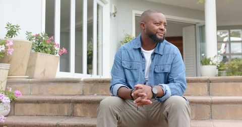 African man sitting on porch steps wearing denim jacket and khaki pants, contemplative