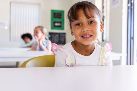 Smiling schoolgirl sitting at front desk in bright classroom with blurred classmates