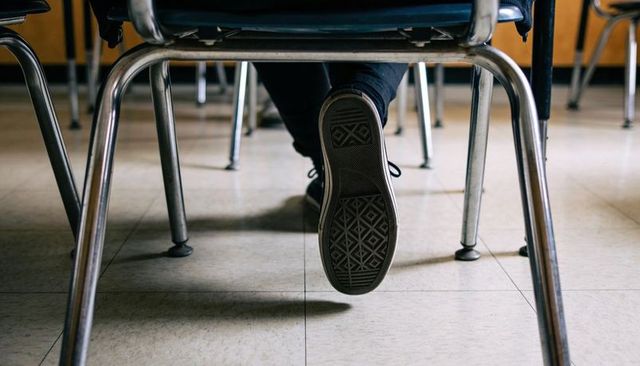 Student dangling sneaker under desk showing sole on classroom tile floor perspective