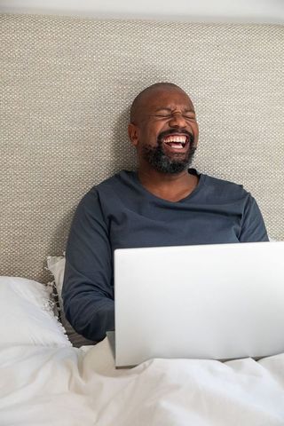 Mature african american man laughing on bed using laptop for remote work and relaxation