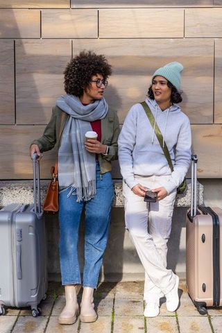 Female friends standing and chatting with carry-on luggage on urban sidewalk