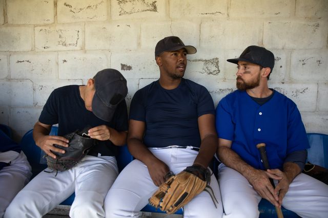 Multi ethnic team of male baseball players before a game, sitting in changing room, interacting, holding bat and glove at a playing field. Baseball sports competition.
