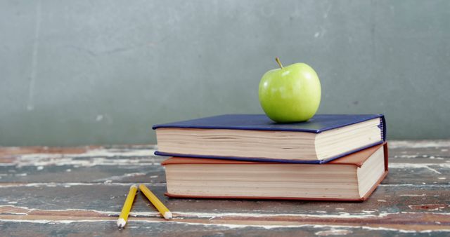 Green apple placed on top of two books on worn wooden desk with pencils beside. Ideal for back to school themes, education concepts, classroom decoration ideas, or study tips illustrations.