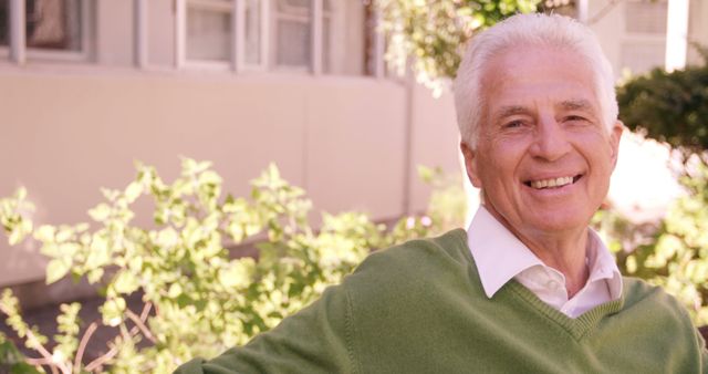 Senior man standing outdoors wearing green sweater and white shirt, smiling in sunlight with greenery in the background. Ideal for use in advertisements or articles about healthy aging, senior lifestyle, positive mindset, retirement, and wellness.