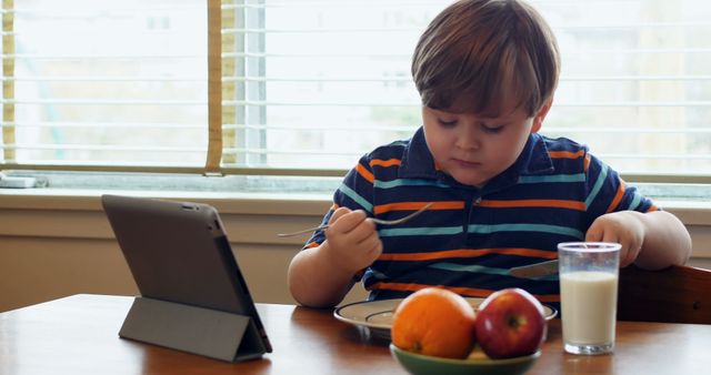Young boy in a striped shirt, sitting at a table, having breakfast with a tablet in front. Add versatile imagery representing technology interwoven with daily routines, youth engaged in screen time, and healthy eating for educational or dietary content. Suitable for articles on children's nutrition, balanced diets, technology in childhood, or family life at home.