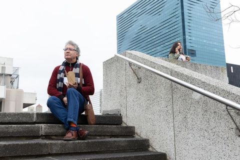 Senior man eating takeaway on city steps while woman talks on phone by glass tower