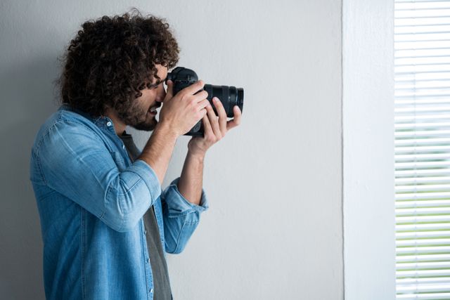 Male photographer with curly hair using a digital camera in a studio. He is wearing a denim shirt and focusing on his subject. This image is ideal for articles or advertisements related to professional photography, creative workspaces, photography tutorials, and equipment reviews.