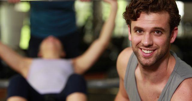 Personal trainer happy in gym while female client working out on bench press in the background. Suitable for fitness training websites, exercise equipment advertisements, or articles on personal training and health.