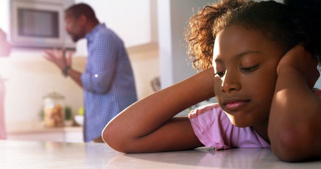 A young African American girl appears sad or contemplative while an adult male, her father, is in the background with a concerned expression, with copy space. Their body language suggests a moment of family tension or a serious conversation taking place.