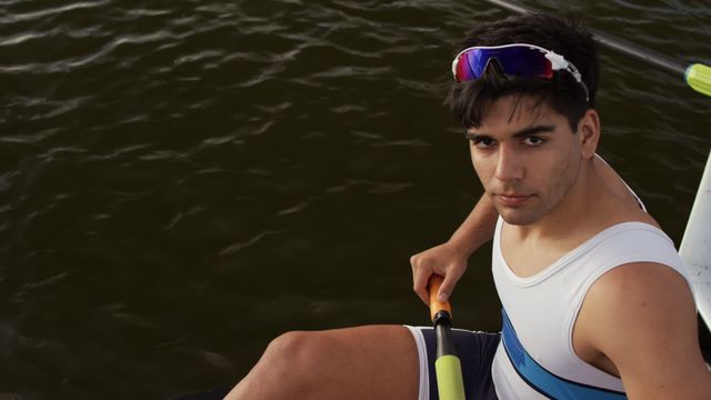 Young male rower taking a break in his boat on the water while looking up at the camera. Suitable for promotions related to sports, outdoor activities and fitness training. Perfect visual for articles about rowing, athlete profiles, or competitive spirit.