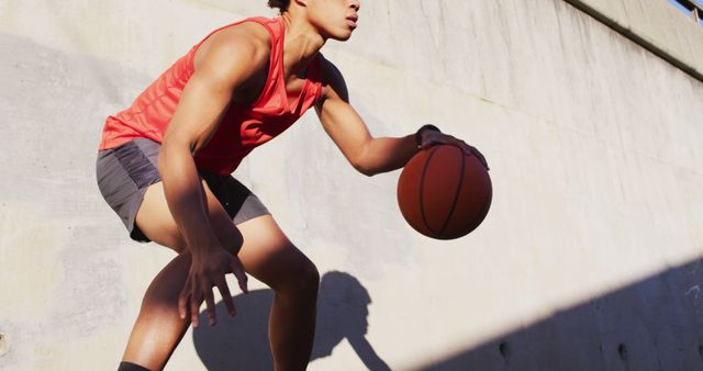 The image shows a young man wearing a red tank top and black shorts dribbling a basketball against a concrete background on a sunny day. The bright light casts clear shadows. This image can be used for promoting sports and fitness activities, illustrating active lifestyles, or in advertisements for sports apparel and equipment.