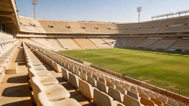Sunlit empty football stadium interior with plastic seats, terraced bleachers and marked green pitch