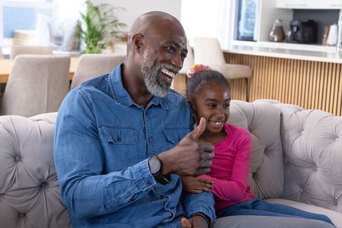 African american grandfather giving thumbs-up while laughing with granddaughter on cozy sofa