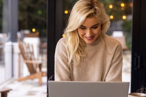Smiling young woman working remotely on laptop in cozy sweater by glass doors with bokeh