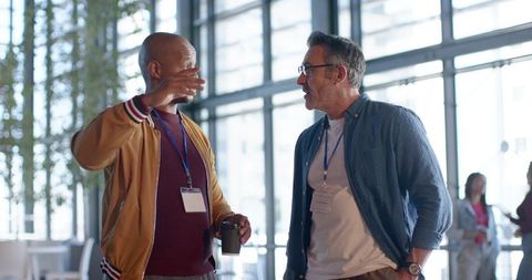 Two male professionals chatting and networking in bright conference lobby holding coffee and badges