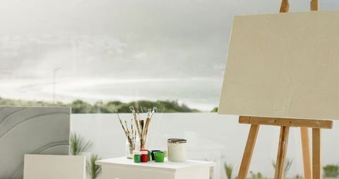 Easel facing misty coastal view with blank canvas, brushes and paint jars