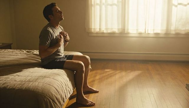 Man sitting on bed holding chest in sunlit minimalist bedroom morning reflection