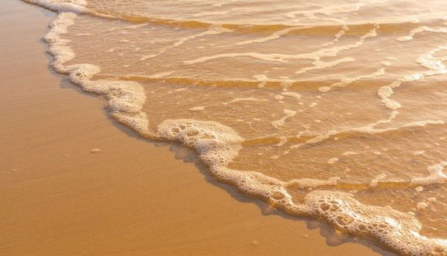 Golden waves lapping sandy shoreline with scalloped foam bubbles at sunset glow