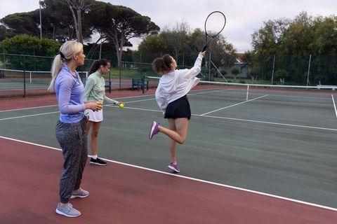 Women practicing tennis on outdoor court, coach guiding players jumping and swinging racket