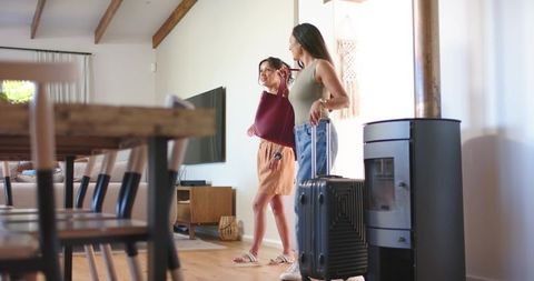 Young women arriving at sunlit vacation rental with suitcase and open-plan living room