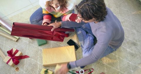 Couple sitting on carpet wrapping gifts with shiny red and gold paper for holiday giving
