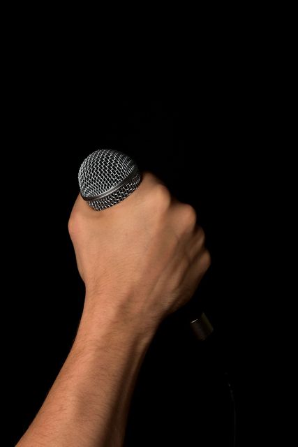 Hand holding microphone tightly against a black background. Ideal for use in presentations, seminars, advertisements for musical equipment, concert posters, public speaking events, and any content related to communication or performance arts.