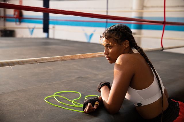 Biracial female boxer practicing in a boxing gym wearing sports clothes, resting next to skipping rope in boxing ring. Strength sports achievement.