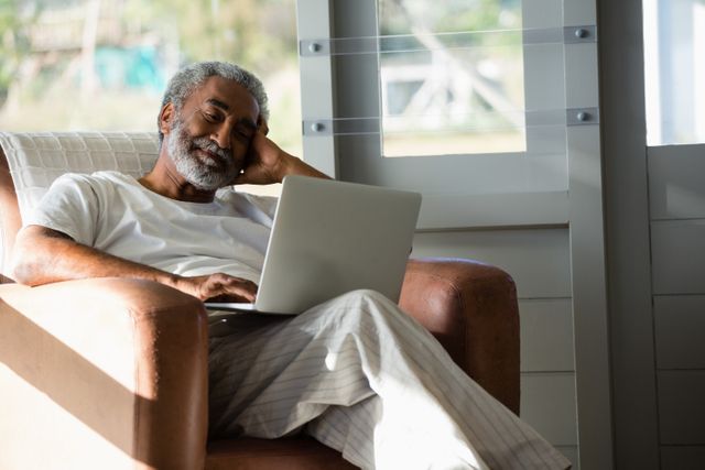 Senior man sitting comfortably in a chair using a laptop at home. He appears relaxed and content, suggesting a leisurely or casual activity. Ideal for use in articles or advertisements about senior lifestyle, technology use among the elderly, retirement activities, or home comfort.