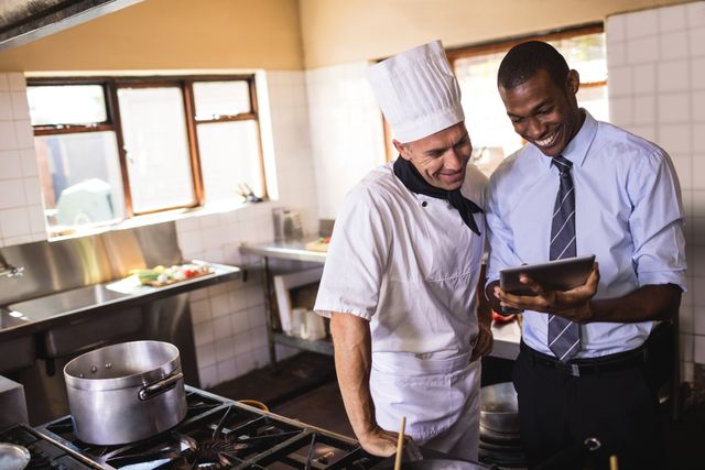 Manager and chef collaborating in a hotel kitchen, using a digital tablet for planning and communication. Ideal for illustrating teamwork, professional environments, and the integration of technology in the hospitality industry. Suitable for use in articles, advertisements, and promotional materials related to hotels, restaurants, and culinary arts.
